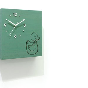 May include: A green wooden clock with white hands and black numbers. The clock face has a black outline of a duck with a bow. The clock is mounted on a white wall.