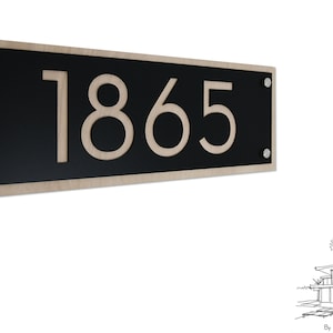 Könnte beinhalten: Ein schwarzes und beiges Holzschild mit der Hausnummer 1865 in beiger Schrift. Das Schild hat auf jeder Seite zwei silberne Schrauben.