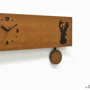 May include: A wooden wall clock with a pendulum and a black silhouette of a deer head. The clock face has black Roman numeral markings and black hands.