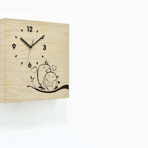 May include: A wooden wall clock with a black clock face and black hands. The clock face has black Roman numerals. The clock has a design of two birds with hearts on a branch. The clock is made by Time Box.