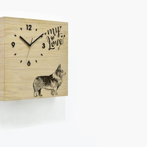 Puede incluir: Reloj de madera con una ilustración en blanco y negro de un perro corgi. La esfera del reloj tiene agujas negras y números negros. El texto "my love" está grabado en la esfera del reloj con corazones.