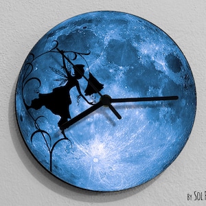 Puede incluir: Un reloj de luna azul con una silueta de un hada balanceándose en una vid. La esfera del reloj es un primer plano de la superficie de la luna. Las manecillas del reloj son negras. El texto "By SOL PIXIE DUST" está en la parte inferior de la imagen.