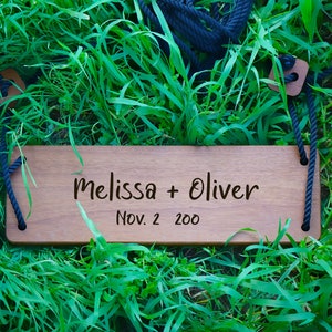 May include: A wooden swing seat with the text "Melissa + Oliver Nov. 2 200" engraved on it. The swing seat is hanging from black ropes and is resting on green grass.