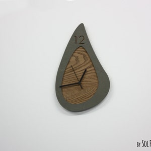 May include: A gray and wood clock with a unique teardrop shape. The clock face is made of wood with a gray border. The clock hands are black and the number 12 is visible on the clock face.