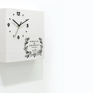 May include: White square wooden clock with black hands and numbers. The clock face has a floral design with two birds and the text "Amelia & Charles 18.03.2019".