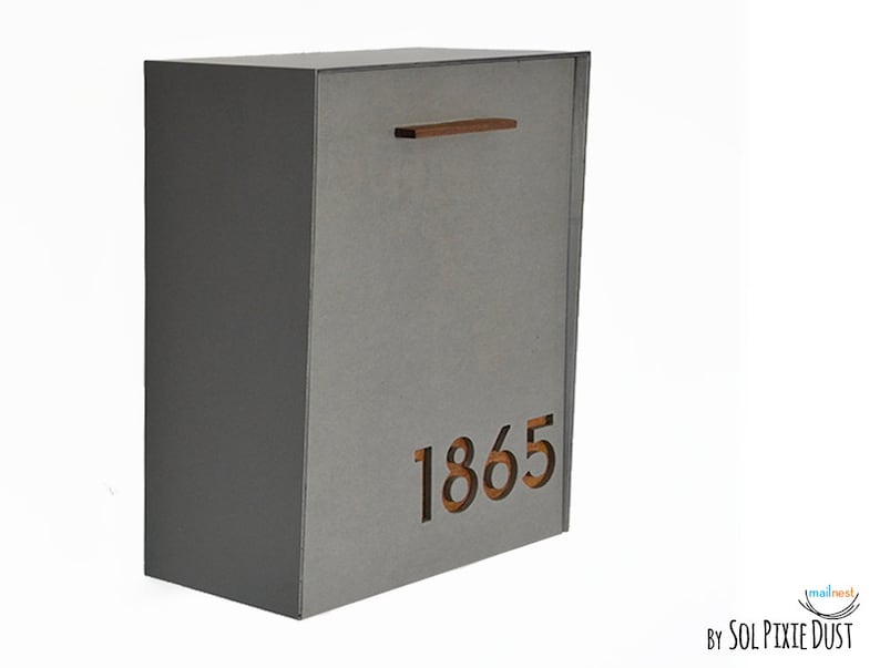 May include: Gray metal mailbox with a wooden door and a cutout of the number 1865. The mailbox is mounted on a white wall.