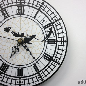 May include: A black and white clock face with Roman numerals and a silhouette of Peter Pan and Tinkerbell flying. The clock face is surrounded by a black border with a repeating pattern of black lines and crosses. The text "By SOL PIXIE DUST" is visible in the bottom right corner of the image.