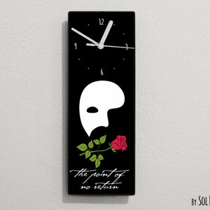 May include: A black rectangular clock with a white face and hands. The clock features a white Phantom of the Opera mask with a red rose and green leaves. The text "the point of no return" is printed below the mask.