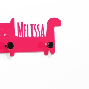 Puede incluir: Un gancho de pared de color rosa en forma de gato con el nombre "Melissa" recortado. El gancho tiene dos ganchos plateados en la parte inferior.