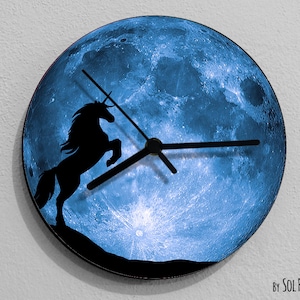 Puede incluir: Un reloj de luna azul con una silueta negra de un unicornio saltando frente a él. Las manecillas del reloj son negras.