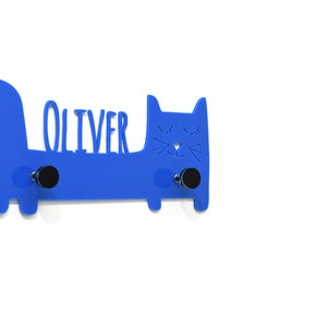 Puede incluir: Un gancho de pared azul en forma de gato con el nombre "Oliver" recortado. El gancho tiene dos ganchos de metal negro.