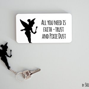 Könnte beinhalten: Ein schwarz-weißer Schlüsselanhänger mit einer Silhouette von Tinkerbell und dem Text "Alles, was du brauchst, ist Glaube - Vertrauen und Feenstaub".