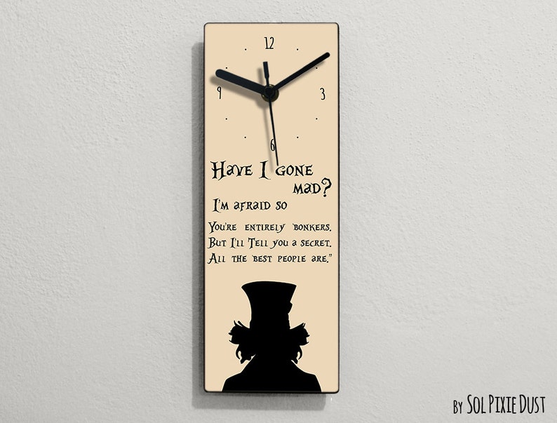 Puede incluir: Un reloj de color crema con agujas negras y una silueta negra del Sombrerero Loco de Alicia en el Pa&iacute;s de las Maravillas. La esfera del reloj tiene la cita "Have I gone mad? I'm afraid so. You're entirely bonkers. But I'll tell you a secret. All the best people are."
