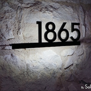 Puede incluir: Placa de número de casa de metal negro con el número 1865 en una pared de piedra blanca.