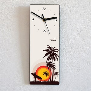 May include: A white clock with black hands and black numbers. The clock face has a sunset scene with palm trees and a surfer. The clock is framed in black.