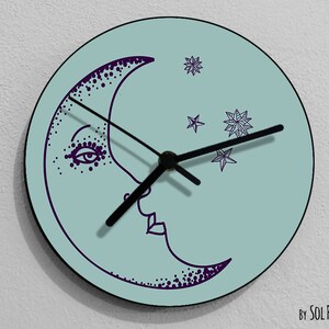 Puede incluir: Un reloj redondo azul claro con manecillas negras y un diseño de luna creciente púrpura con estrellas. La luna tiene una cara con un ojo y una boca. El texto "By SOL PIXIE DUST" está en la parte inferior del reloj.