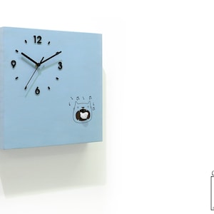 May include: A light blue square wall clock with black hands and numbers. The clock features a cartoon cat singing with musical notes. The clock is made by Time Box, a Cuckoo Clocks brand.