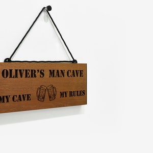 May include: A wooden sign with the text "Oliver's Man Cave My Cave My Rules" and a graphic of two beer mugs clinking. The sign is hanging from a black cord.