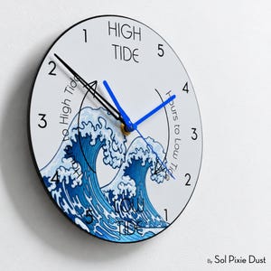 Op de afbeelding: Een ronde wandklok met een witte wijzerplaat en een blauwe illustratie van een oceaangolf. De klok toont de tekst HIGH TIDE en LOW TIDE, met cijfers 1 tot 5 die de uren tot vloed en eb aangeven. De klok heeft zwarte en blauwe wijzers.