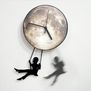 Puede incluir: Un reloj con forma de luna con agujas negras y fondo blanco. Dos siluetas negras de personas se balancean en los brazos del reloj.