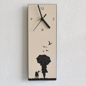 Könnte beinhalten: Eine beige Wanduhr mit schwarzen Zeigern und Zahlen. Das Zifferblatt zeigt eine Silhouette einer Person, die einen Hund mit einem Regenschirm spazieren führt, und Vögel, die über ihnen fliegen.