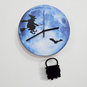 Può includere: Un orologio blu e bianco con una strega che vola su una scopa e un pipistrello. L'orologio ha un pendolo nero a forma di calderone.