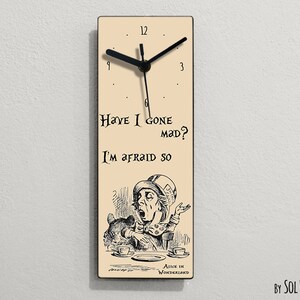Puede incluir: Un reloj de madera beige con agujas negras y n&uacute;meros romanos negros. La esfera del reloj presenta una ilustraci&oacute;n en blanco y negro del Sombrerero Loco de Alicia en el Pa&iacute;s de las Maravillas, con el texto "Have I gone mad? I'm afraid so." y "Alice in Wonderland".
