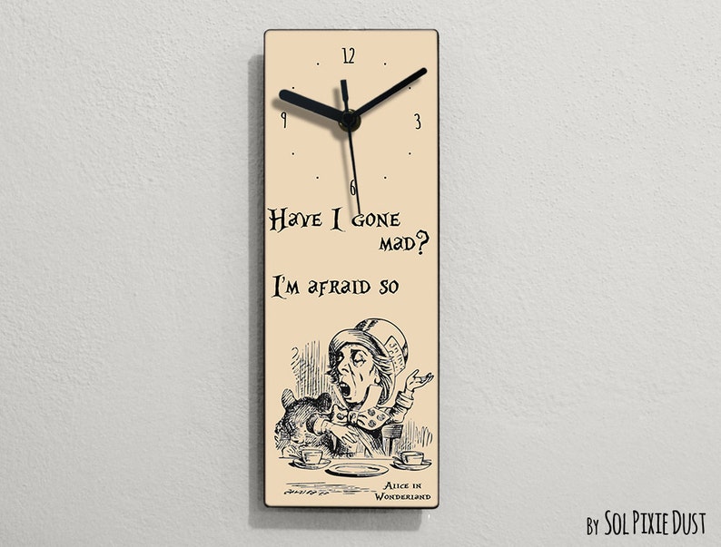 Puede incluir: Un reloj de madera beige con agujas negras y n&uacute;meros romanos negros. La esfera del reloj presenta una ilustraci&oacute;n en blanco y negro del Sombrerero Loco de Alicia en el Pa&iacute;s de las Maravillas, con el texto "Have I gone mad? I'm afraid so." y "Alice in Wonderland".