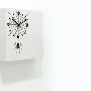 May include: A white square clock with a black spider web and a black spider hanging from the bottom. The clock face has black numbers and hands.