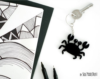 May include: Black crab-shaped keychain with a silver key ring and a silver key attached. The keychain is on a white background with a black and white drawing and black markers.