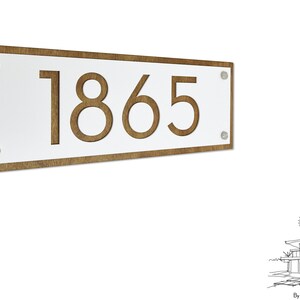 Könnte beinhalten: Ein rechteckiges weißes Hausnummernschild mit der Nummer 1865, die aus braunem Holz ausgeschnitten ist. Das Schild ist auf einem braunen Holzrahmen montiert und hat auf jeder Seite zwei silberne Schrauben.