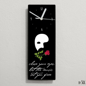 May include: Black rectangular clock with a white clock face and hands. The clock has a white phantom mask with a red rose in its mouth. The text "close your eyes, let the music set you free" is below the mask. The clock is on a white wall.