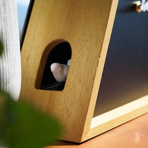 May include: A wooden clock with a black face and a small white bird figurine peeking out of a cutout in the wood. The clock is on a wooden surface.