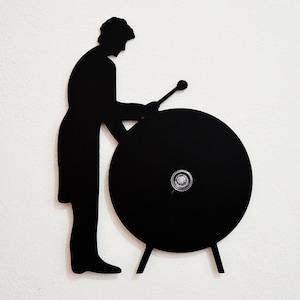 May include: Black silhouette of a person playing a large gong with a mallet. The gong is round and has a small knob in the center.