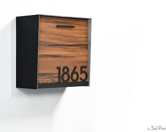 Puede incluir: Un buzón de pared con marco de metal negro y panel frontal con veta de madera. El panel frontal incluye un asa negra horizontal en la parte superior y los números de casa negros 1865 situados en la esquina inferior derecha.