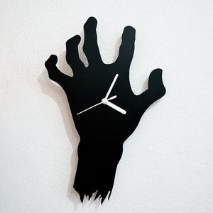 Op de afbeelding: Zwarte wandklok in de vorm van een uitgestrekte hand. De klok heeft witte wijzers en een eenvoudig ontwerp.