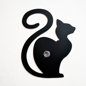 May include: Black metal silhouette of a cat with its tail curled around its body. The cat is mounted on a white wall.