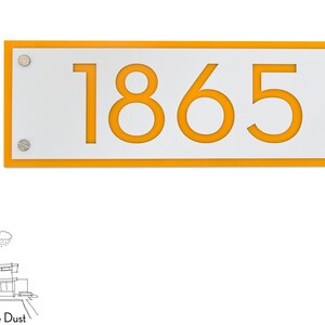 Könnte beinhalten: Ein rechteckiges Hausnummernschild aus weißem Material mit orangefarbenem Rand und der Nummer 1865 in Orange.