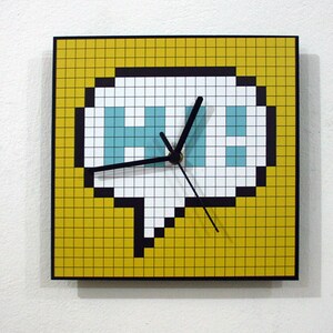 Op de afbeelding: Een gele vierkante klok met een zwarte rand. De wijzerplaat van de klok is een witte gepixelde spraakballon met het woord "HI" in blauwe pixels. De wijzers van de klok zijn zwart.