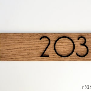 Op de afbeelding: Een houten bord met het nummer 203 in zwart gedrukt. Het bord is gemaakt van lichtbruin hout met een natuurlijke houtnerf. De tekst "By Sol Pixie Dust" is in zwart gedrukt aan de onderkant van het bord.