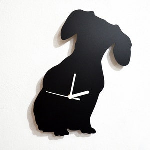 Puede incluir: Un reloj de pared negro con la forma de un perro salchicha. El reloj tiene manecillas blancas y un diseño sencillo y moderno. La silueta del perro está contra una pared blanca.