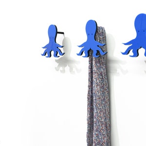 Może przedstawiać: Trzy niebieskie haczyki ścienne w kształcie ośmiornicy, jeden z wiszącą wzorzystą szalikiem. Haczyki są zamontowane na białej ścianie.