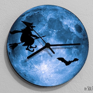 Puede incluir: Un reloj de luna azul con una silueta negra de una bruja volando en una escoba y un murciélago. Las manecillas del reloj son negras. El texto "By SOL PIXIE DUST" está en la parte inferior del reloj.