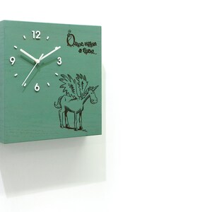 May include: A square clock with white hands and black numbers, set against a green background. The clock face features a black and white illustration of a winged unicorn and the text "Once upon a time..."