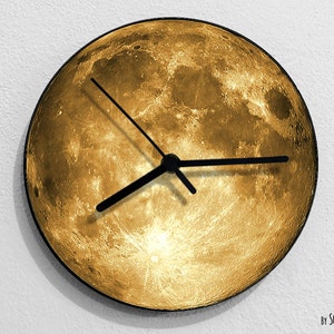Puede incluir: Un reloj de color dorado con agujas negras, que presenta una imagen de luna llena en la esfera. El reloj está montado en una pared blanca. El texto "By SOL PIXIE DUST" es visible en la parte inferior de la imagen.