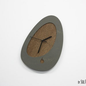 May include: A gray, pebble-shaped wall clock with a wood-grain face and black hands. The clock is hanging on a white wall.