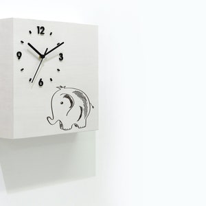 May include: White square clock with black hands and numbers. The clock has a black and white illustration of an elephant below the clock face. The clock is mounted on a white wall. The text "Time Box" is in the lower right corner of the image.