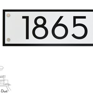 Könnte beinhalten: Ein rechteckiges weißes Hausnummernschild mit schwarzem Rand und schwarzen Zahlen. Das Schild zeigt die Nummer "1865". Das Schild ist mit silbernen Schrauben befestigt.