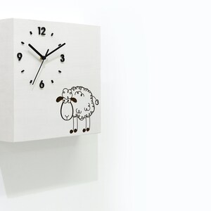 May include: White square clock with black hands and numbers. The clock has a black and white cartoon sheep illustration below the clock face. The clock is mounted on a white wall.