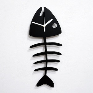 May include: Black fish-shaped wall clock with white hands and a skeleton design. The clock is mounted on a white wall.